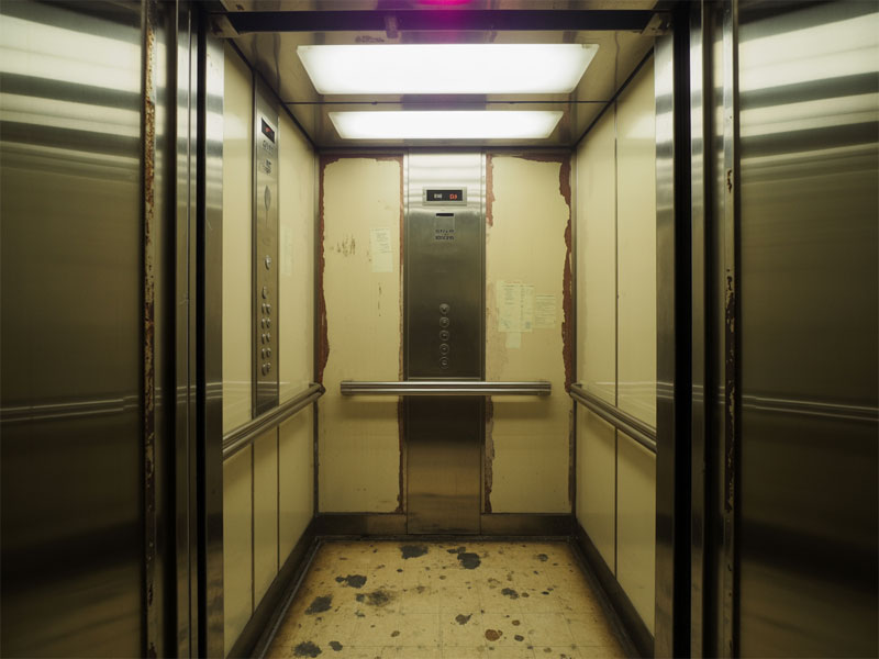 An old, worn elevator interior with peeling walls, visible stains on the floor, and a central control panel under fluorescent lighting.